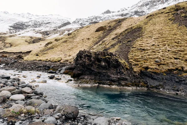 Rio de montanha no sopé da montanha com um pico coberto de neve. Grama seca amarela nas montanhas em maio na Islândia . — Fotografia de Stock