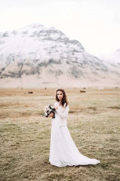 Potret seorang pengantin dengan gaun pengantin putih, dengan buket pengantin di tangannya. Di lapangan rumput kuning kering, di tengah gunung bersalju dan merumput kuda Islandia. Tujuan pernikahan Islandia.. — Stok Foto