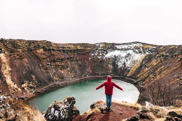 A guy in a red jacket stands on a rock above Lake Kerid - a crater volcanic lake in Iceland. Red volcanic soil, similar to Martian landscapes. Hands thrown upside down and rejoices.