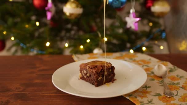 Un morceau carré de Brownie au chocolat sur une assiette, sur une table en bois arrosé de caramel salé à ficelle dorée, il s'égoutte sur les bords du gâteau. Dans le contexte de l'arbre de Noël décoré . — Video