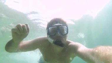 Güzel genç adam dalış maskesiyle şnorkelle yüzüyor ve denizde berrak mavi suda yüzerken kameraya el sallıyor. Balıklı ve mercan resifli sualtı dünyası. Adam özçekim yapıyor.