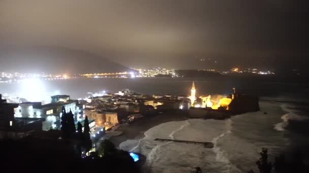 Onde di tempesta sulla spiaggia vicino al centro storico di Budva, Montenegro. La luce della città di notte . — Video Stock