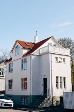 Kırmızı çatılı beyaz dikdörtgen bir evin ön cephesi, İzlanda 'nın başkenti Reykjavik' te yol boyunca parçalanmış beyaz bir araba..