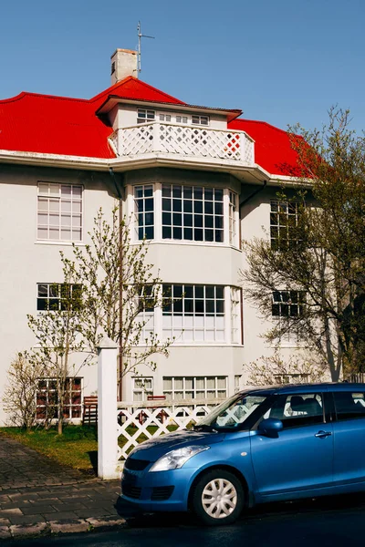 Maison de trois étages avec de grandes fenêtres panoramiques et un grenier au toit rouge à Reykjavik, la capitale de l'Islande. Une voiture bleue est garée près de la maison le long de la route . — Photo