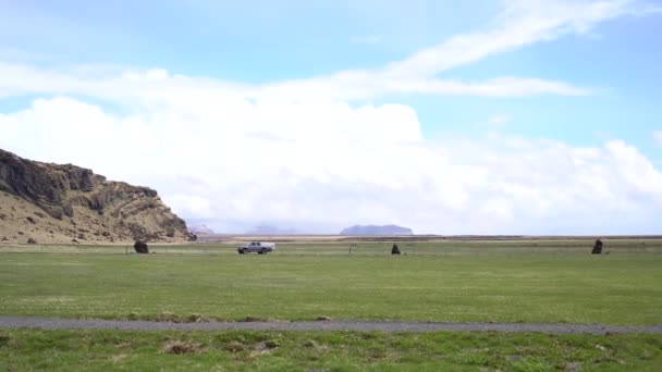 Um caminhão pickup passeios ao longo da estrada, entre a grama verde, contra o pano de fundo de montanhas e céus azuis com nuvens brancas na Islândia . — Vídeo de Stock