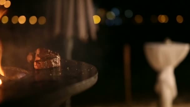 Gros plan du pain de viande sur un grill en métal, mouvement de camée, lumières de la ville nocturne. Steak de viande est en cours de préparation sur un barbecue extérieur en acier rond avec une table de cuisson et un feu ouvert au milieu .