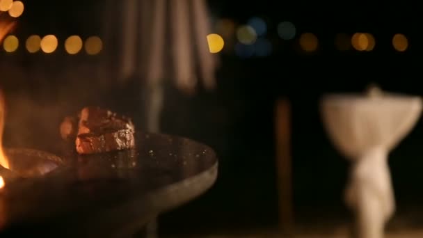 Close-up do pão de carne em uma grade de metal, movimento de cameo, luzes da cidade noturna. Bife de carne está sendo preparado em uma grade ao ar livre de ferro de aço redondo com uma placa de cozinhar e uma fogueira aberta no meio . — Vídeo de Stock
