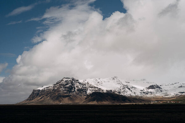 A wide field of dry yellow grass and a mountain in the distance with a snow-capped peak, against a blue sky with white clouds in Iceland.