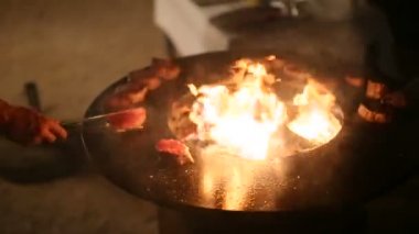 Şef kızartma makinesinde biftekleri maşayla çeviriyor. Et bifteği çelik bir ızgarada pişiriliyor. Bir tenceresi ve ortasında açık bir ateş var..