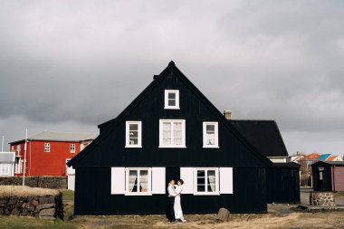 Varış yeri İzlanda düğünü. Beyaz pencereli ve panjurlu siyah ahşap bir evin yanında düğün çifti. Damat gelini kucaklıyor..