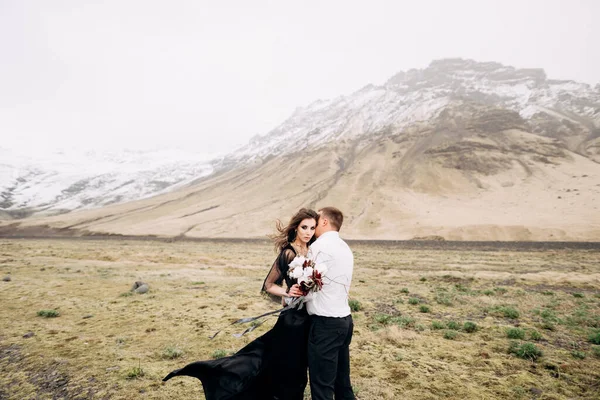 Casamento casal em um fundo de montanhas nevadas. A noiva em um vestido preto e noivo estão abraçando em um campo de musgo e grama amarela. Destino Islândia casamento . — Fotografia de Stock