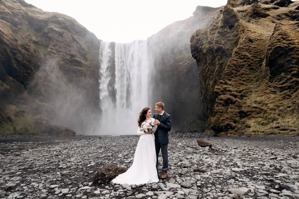 Pasangan pernikahan dekat air terjun Skogafoss. Tujuan pernikahan Islandia. Pengantin pria memeluk mempelai wanita dengan bahu, mereka tertawa bersama. — Stok Foto