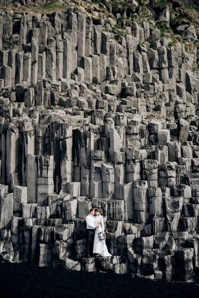 Un couple de mariés se tient sur un mur de piliers de pierre. Les mariés s'étreignent sur le basalte Kekurs, sur la plage de sable noir de Vik. Destination Mariage Islande . — Photo
