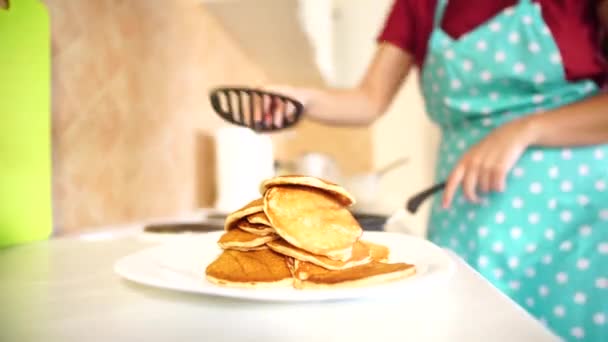 Time-lapse The girl in the blue apron cooks pancakes. A stack of pancakes in the foreground. Removes the finished pancakes from the pan and pours a ladle a new batch. — Stock Video