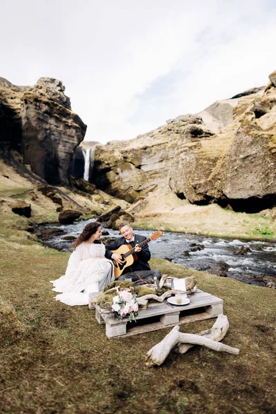 Um casal de casamentos está sentado na margem de um rio de montanha, em uma mesa para um jantar de casamento. O noivo toca e canta para a noiva. Casamento destino Islândia, perto de cachoeira Kvernufoss . — Fotografia de Stock