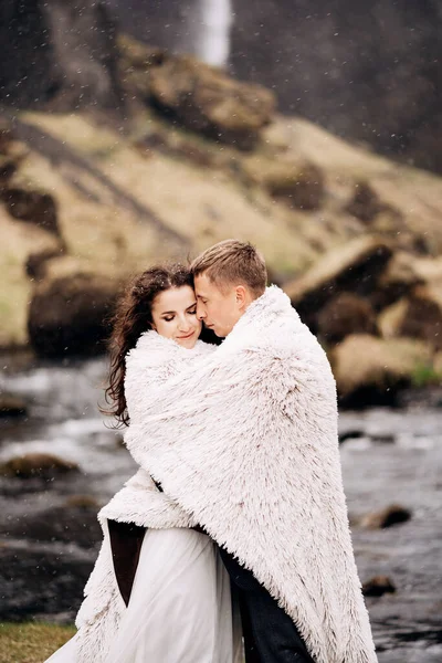 Um casal de casamentos fica na margem de um rio de montanha sob um cobertor de lã. O noivo abraça a noiva, tentando aquecê-la. A neve cai. Casamento destino Islândia, perto de cachoeira Kvernufoss . — Fotografia de Stock