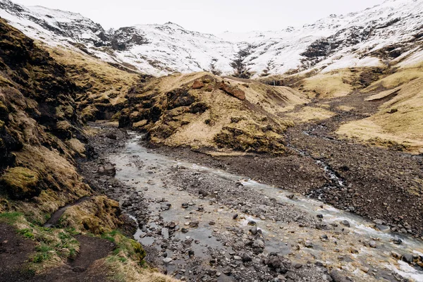 Hegyi folyó a hegy lábánál, hófödte csúccsal. Sárga száraz fű a hegyekben májusban Izlandon. — Stock Fotó