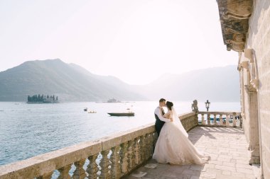 Bir düğün çifti, eski Perast kasabasında günbatımında Kotor Körfezi ve St. George ve Gospa od Skrpela adasına bakan taş sütunlar ve aslan heykelleriyle büyük bir terasta duruyor..