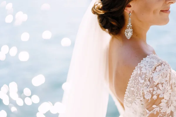 Close-up of the neck and neck of the bride, face profile. Beautiful big earring in ears, shoulder with white lace dress on background of glare of water. Fine-art wedding photo in Montenegro, Perast. — Stock Photo, Image