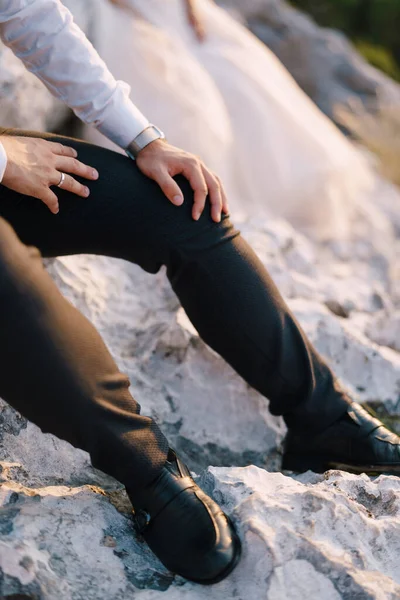 Fine-art destination wedding photo in Montenegro, Mount Lovchen. Hands of the groom with a watch and a ring lie on their knees, black shoes and dark blue trousers on their feet. — Stock Photo, Image