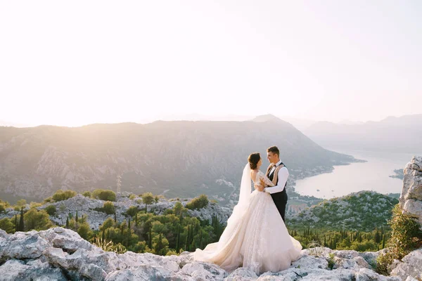 Ein Hochzeitspaar steht auf einem Berg mit Panoramablick auf die Bucht von Kotor bei Sonnenuntergang. Der Bräutigam umarmt die Braut sanft. Hochzeitsfoto in Montenegro, Berg Lovchen. — Stockfoto