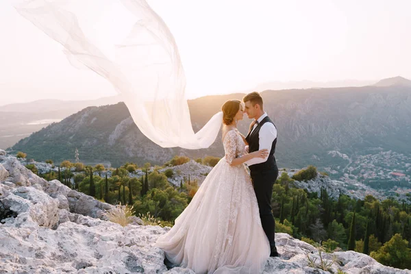 Fine-Art-Reiseziel Hochzeitsfoto in Montenegro, Mount Lovchen. Das Hochzeitspaar umarmt sich bei Sonnenuntergang auf dem Gipfel des Berges, der Bräutigam umarmt die Braut an der Taille, der Schleier flattert in die Luft. — Stockfoto