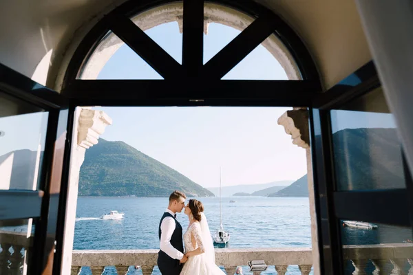 Nevěsta a ženich stojí na balkoně hotelu s výhledem na moře, pohled otevřeným starým oknem. Luxusní svatební fotografie v Černé Hoře, Perast. — Stock fotografie