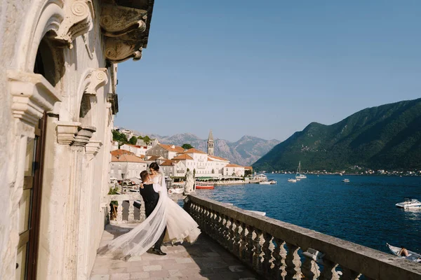 Foto de casamento de arte em Montenegro, Perast. Um casal de casamentos no terraço do hotel com vistas panorâmicas da cidade velha de Perast, o noivo circunda a noiva com um vestido longo em suas mãos . — Fotografia de Stock