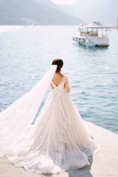 A menyasszony áll a mólón a város Perast Montenegróban, és megcsodálja a hegyek és a kikötött hajók. A ruha vonata a padlón fekszik, és hosszú fátyol tör ki a szélben.. — Stock Fotó