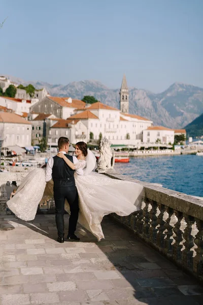 Foto de casamento de arte em Montenegro, Perast. Um casal de casamentos no terraço do hotel com vistas panorâmicas da cidade velha de Perast, o noivo circunda a noiva com um vestido longo em suas mãos . — Fotografia de Stock