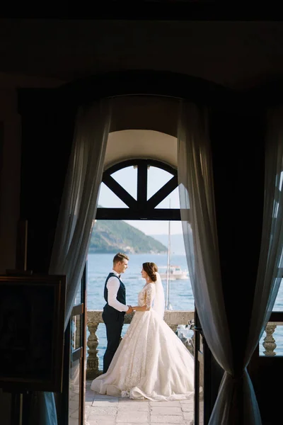 Bruden och brudgummen står på balkongen på hotellet med utsikt över havet, en titt genom det öppna gamla fönstret. Fine-art bröllop foto i Montenegro, Perast. — Stockfoto