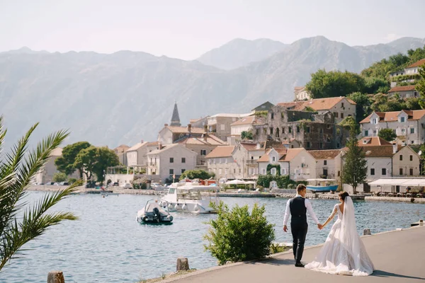 Foto de casamento de arte em Montenegro, Perast. Um casal de casamentos caminha ao longo do calçadão perto do mar, em meio a palmeiras, montanhas e a cidade velha . — Fotografia de Stock