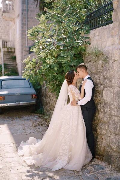 Pasangan pengantin berpelukan di jalan-jalan kota tua, di bawah pohon ara, dengan latar belakang mobil Yugoslavia tua. Foto pernikahan seni rupa di Montenegro, Perast. — Stok Foto