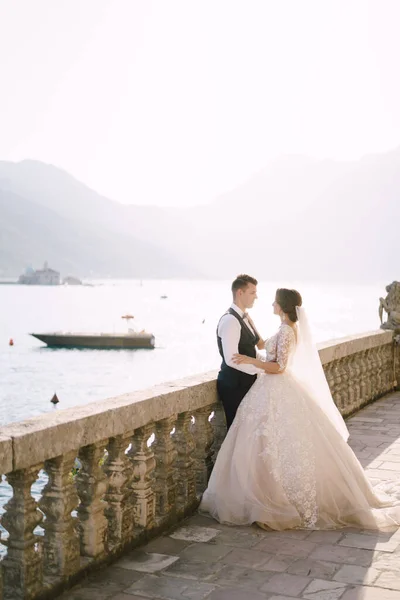 A noiva e o noivo estão em pé em um grande terraço com colunas de pedra, com vista para a Baía de Kotor e a ilha de São Jorge e a Ilha de Gospa od Skrpela ao pôr do sol na cidade velha de Perast — Fotografia de Stock