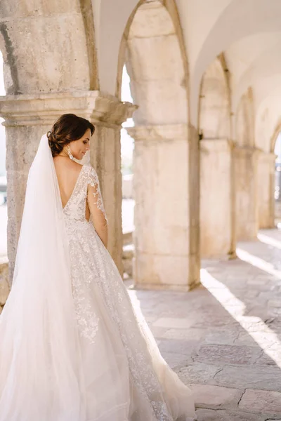 Seorang pengantin dalam gaun putih dan dengan kerudung panjang berdiri dengan punggungnya ke kamera, dalam kolom batu putih melengkung. Foto pernikahan seni rupa di Montenegro, Perast. — Stok Foto