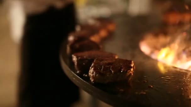 En närbild av squaring kött på den heta ytan. Köttbiff förbereds på en rund utegrill av järn med spishäll och öppen eld i mitten. — Stockvideo