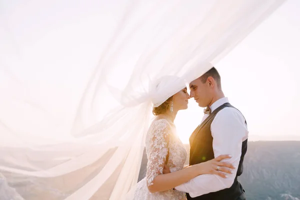 Foto de casamento de destino de arte em Montenegro, Monte Lovchen. Os abraços de casal de casamento no topo da montanha ao pôr do sol, o noivo abraça a noiva pela cintura, o véu ondulado no ar . — Fotografia de Stock