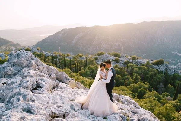 Um casal de casamentos fica no topo de uma montanha com vistas panorâmicas da Baía de Kotor, ao pôr-do-sol. O noivo gentilmente abraça a noiva. Fotos de casamento de destino de arte em Montenegro, Monte Lovchen . — Fotografia de Stock