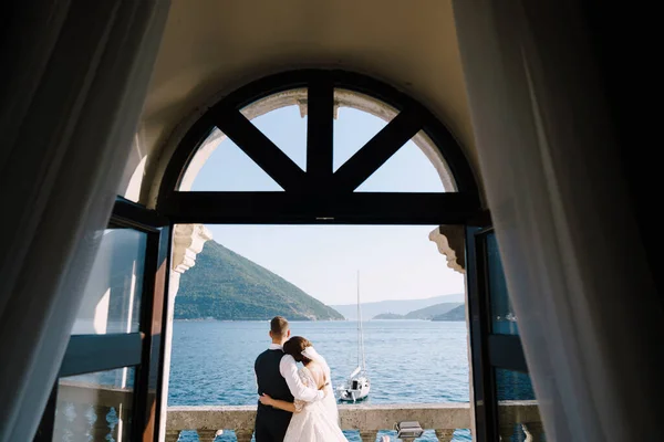 Svatební pár stojí na hotelovém balkóně s výhledem na moře, výhled přes otevřené starožitné okno. Luxusní svatební fotografie v Černé Hoře, Perast. — Stock fotografie