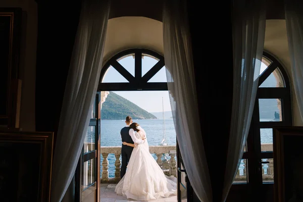 Bröllopspar står på en hotellbalkong med havsutsikt, utsikt genom ett öppet antikt fönster. Fine-art bröllop foto i Montenegro, Perast. — Stockfoto