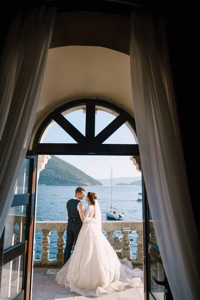 Bröllopspar står på en hotellbalkong med havsutsikt, utsikt genom ett öppet antikt fönster. Fine-art bröllop foto i Montenegro, Perast. — Stockfoto