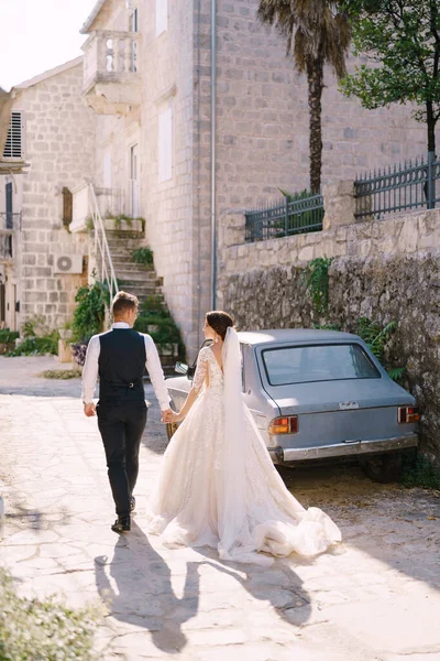 Pengantin wanita dan pengantin pria berjalan di sepanjang jalan batu tua, dengan latar belakang mobil vintage biru. Pasangan pernikahan berpegangan tangan, bayangan di lantai. Foto pernikahan seni rupa di Montenegro, Perast. — Stok Foto