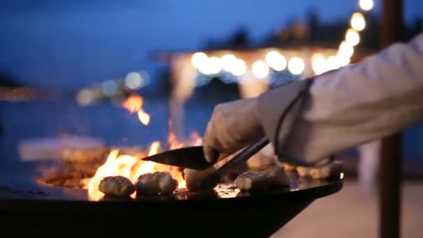 Le chef enlève les steaks de poisson du gril avec une spatule et une pince. Le poisson est en cours de préparation sur un barbecue extérieur en acier rond avec une table de cuisson et un feu ouvert au milieu. — Video