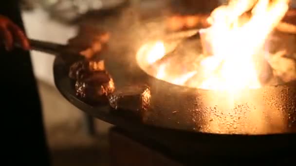 De chef-kok slaat de stukken vlees met een tang, meet de temperatuur in het vlees. Vleesbiefstuk wordt bereid op een ronde stalen buitengrill met een kookplaat en een open vuur in het midden. — Stockvideo