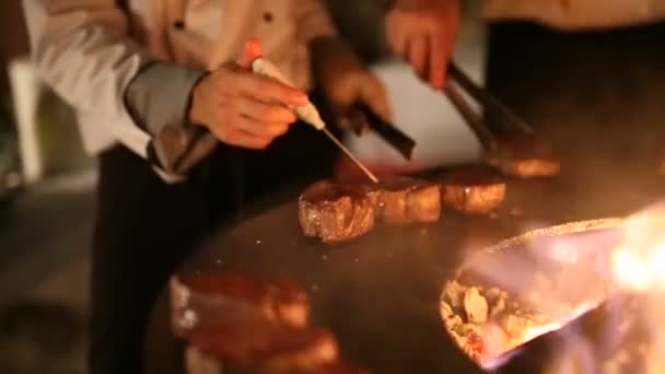 O chef vira os pedaços de carne com fórceps, mede a temperatura dentro da carne. Bife de carne está sendo preparado em uma grade ao ar livre de ferro de aço redondo com uma placa de cozinhar e uma fogueira aberta no meio . — Vídeo de Stock