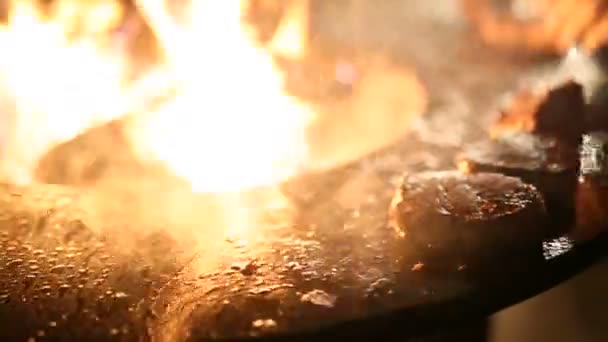 O bife de carne de close-up está sendo preparado em uma grade ao ar livre de ferro de aço redondo com uma placa de fogão e uma lareira aberta no meio . — Vídeo de Stock