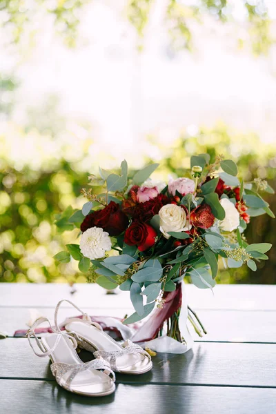 Ramo nupcial de rosas blancas y rojas, peonías, ramas de eucalipto, alstroemeria y crisantemo con cintas blancas y granate, zapatos de la novia cerca de ella —  Fotos de Stock