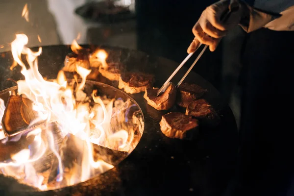 Szef kuchni grilluje steki. Zmienia mięso szczypcami. Grillowany okrągły stół z ogniem wewnątrz. — Zdjęcie stockowe