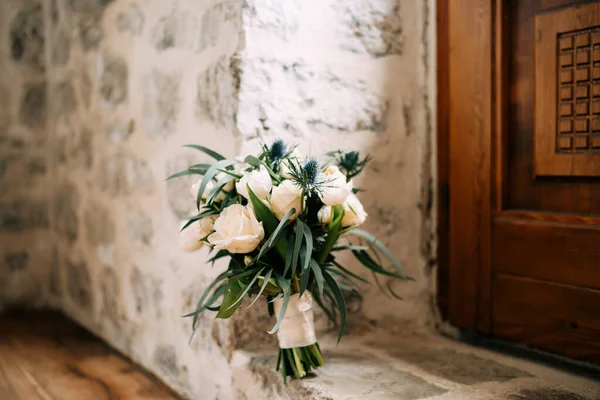 Ramo nupcial de rosas rosadas y blancas, tulipanes, ramas de eucalipto, eryngium, scabiosa y cintas blancas en el suelo cerca de la puerta de madera y la pared de piedra —  Fotos de Stock