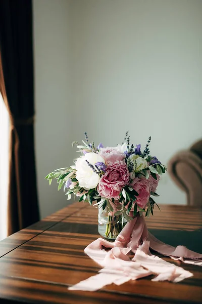 Ramo nupcial de peonías blancas y rosadas, ramas de olivo, lavanda y campanula con cintas rosadas en el jarrón de vidrio sobre la mesa en la habitación —  Fotos de Stock